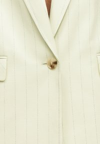 Ανοιχτό μπεζ σακάκι με λεπτές ρίγες, με ένα καφέ κουμπί κλεισμένο και δύο τσέπες με καπάκι, που δείχνει γιακά και άνω μέρος του στήθους.