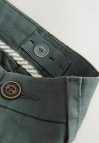 Cintura verde ajustable en pantalones con botón y banda elástica con múltiples ojales para personalizar el ajuste.