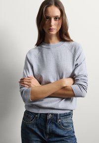 Pull en tricot gris avec un col et des poignets côtelés, présentant une coupe décontractée et une texture douce, associé à un jean en denim bleu.