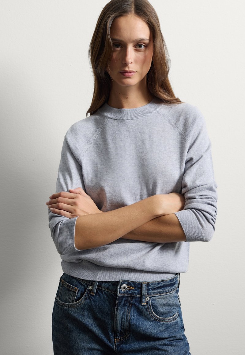 Pull en tricot gris avec un col et des poignets côtelés, présentant une coupe décontractée et une texture douce, associé à un jean en denim bleu.