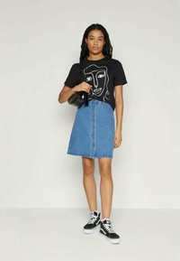 Μαύρο t-shirt με λευκό αφηρημένο γραφικό προσώπου, σε συνδυασμό με μπλε τζιν φούστα με εμπρόσθια κουμπώματα. Το μοντέλο κρατάει μια μαύρη τσάντα.