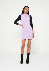 Megzta suknelė su šviesiai levandų spalvos kūnu ir tamsiai mėlynomis ilgomis rankovėmis, aukšta apykakle ir logotipo detalėmis ant krūtinės. Lygi tekstūra.