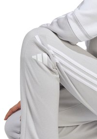 Helle graue Sporthose mit weißem Drei-Streifen-Design, aus weichem Material. Sie hat eine schlanke Passform und ein dezentes Logo-Detail an der Seite.