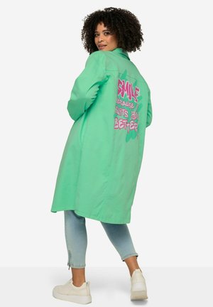 Long mint green jacket made of cotton, featuring a pink graphic text design on the back. Paired with light denim and white sneakers.