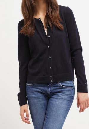 Vrouw draagt een nauwsluitend marineblauw vestje met knopen dicht over een wit shirt, gecombineerd met blauwe spijkerbroek, tegen een effen achtergrond.