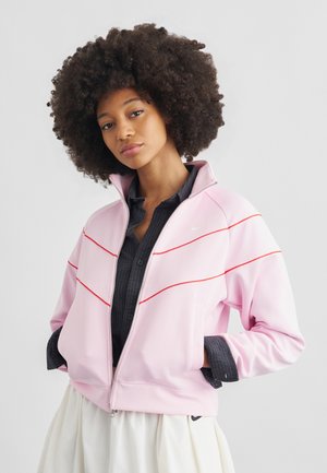 Mlajša ženska s kodrastimi lasmi, oblečena v svetlo roza jakno z rdečimi črtami čez temno srajco in belo krilo, s rokami v žepih.