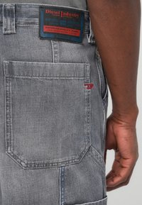 Jeans in denim grigio con una toppa rettangolare sul retro con testo rosso e blu, tasche standard e un dettaglio decorativo di cuciture rosse.