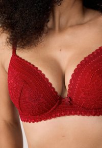Czerwona koronkowa biustonosz z dekoltem w kształcie litery V, falbaniastymi krawędziami i detalem w postaci kokardy na środku, zdobiony kwiatowymi i geometrycznymi wzorami.