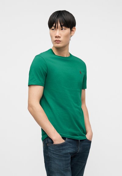 Νέος άνδρας που φοράει πράσινο κοντομάνικο μπλουζάκι και σκούρο τζιν με τα χέρια στις τσέπες, κοιτάζοντας προς τα αριστερά του, μπροστά σε μια απλή φόντο.