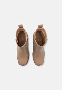 Botines color canela hechos de cuero liso con punta cuadrada, detalle de cremallera en el lateral y una lengüeta para tirar en la parte trasera. Forro interior suave.