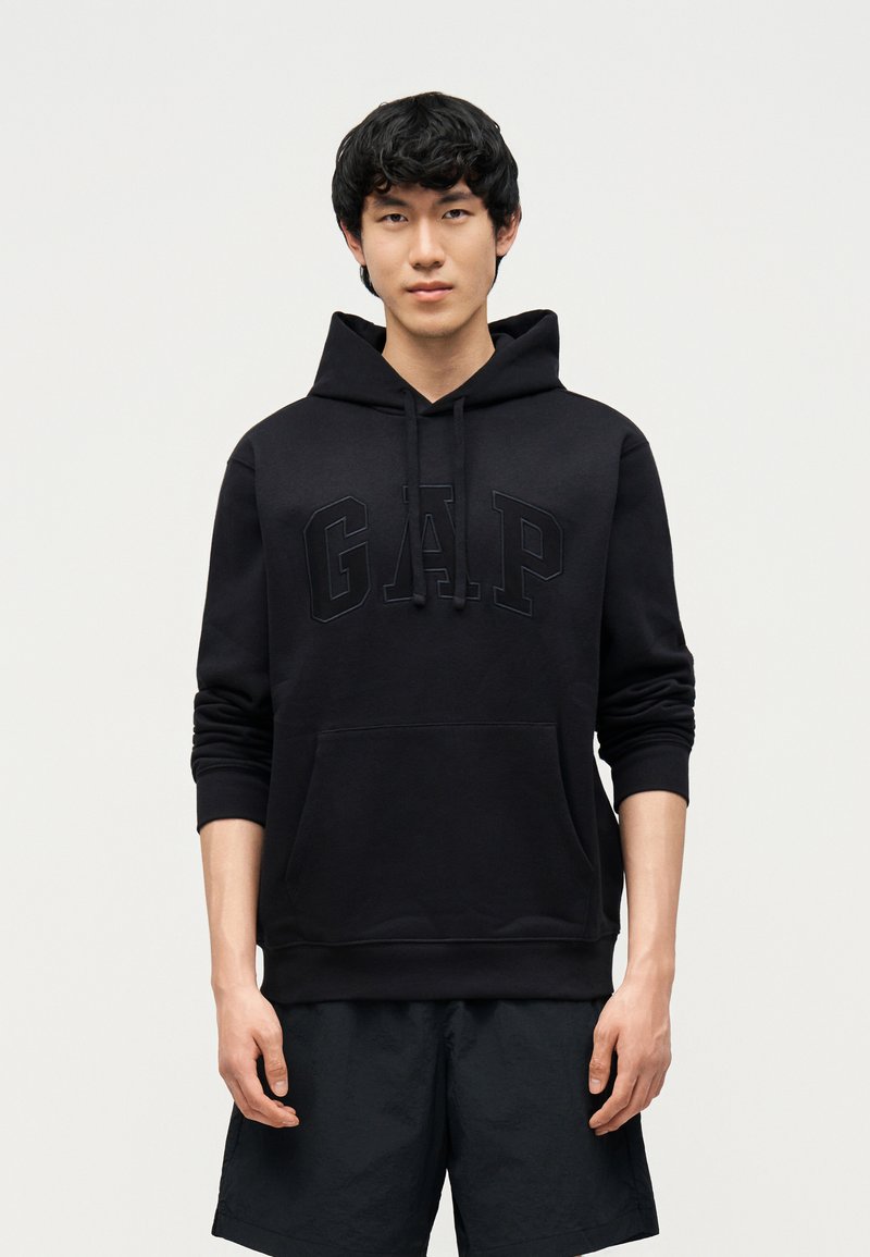 Νεαρός άνδρας που φοράει μαύρο φούτερ με κουκούλα με το ανάγλυφο λογότυπο «GAP» στο μπροστινό μέρος και μαύρο σορτς, στέκεται μπροστά σε ένα απλό ανοιχτό φόντο.