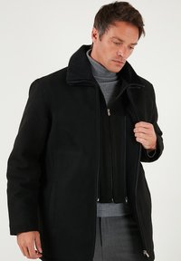 Melnā jaka ar mīkstu, teksturētu apkakli, ar rāvējslēdzēju priekšā un divām sānu kabatām, ģērbta virs pelēka turtlenecka džempera.