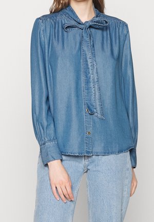 Mujer que lleva una blusa larga de mezclilla azul claro con cuello de lazo y vaqueros de mezclilla azul claro.