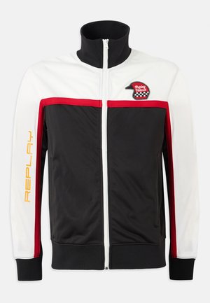 Giacca a pista nera, bianca e rossa con colletto alto, strisce rosse, zip frontale e un logo raffigurante un casco sul lato sinistro del petto. "REPLAY" stampato in giallo.