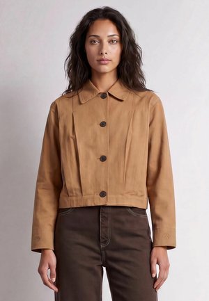Donna con capelli lunghi e scuri che indossa una giacca beige abbottonata e pantaloni marrone scuro, in piedi davanti a uno sfondo chiaro e uniforme.