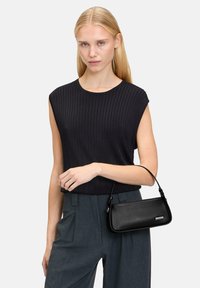 Black leather handbag with a sleek, rectangular shape and a curved handle. Model wears a black ribbed top and gray high-waisted trousers.