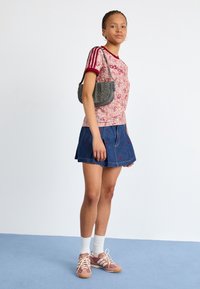 Young woman wearing a floral Adidas t-shirt, denim skirt with red embroidery, white socks, pink sneakers, and a gray studded shoulder bag.