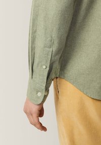 Camisa verde de botones con una tela texturada, con botones blancos y un dobladillo curvado. Lleva puesta sobre pantalones de pana color marrón claro.