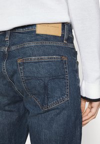 Tmavě modré džíny z denimu s koženou nášivkou s nápisem "TIGER OF SWEDEN" v pase, s oranžovým prošíváním a standardním designem kapes.