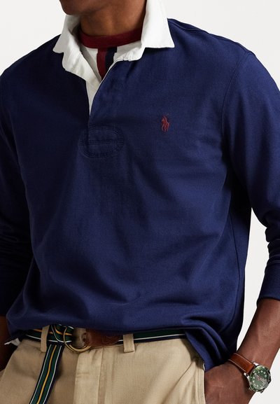 Marineblauwe polo met lange mouwen, voorzien van een witte kraag, een klein logo op de borst en een textuurstof. Riem in gestreept groen en wit.