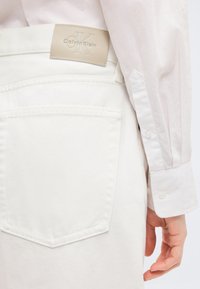 Κοντινό πλάνο της τσέπης λευκού τζιν με μπεζ μπατζάκι λογότυπου Calvin Klein και το χέρι ενός ατόμου να ακουμπά δίπλα της.