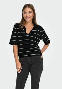 Young woman smiling, wearing a black short-sleeve striped shirt and dark jeans, hand resting on hip, standing against a plain background.