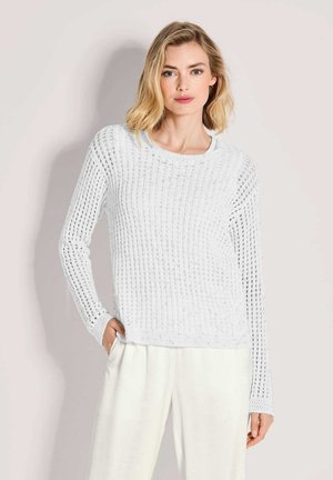 Frau mit blondem Haar, die einen weißen, offen gestrickten Pullover und weite weiße Hosen trägt, mit einer Hand in der Tasche, stehend vor einfachem Hintergrund.