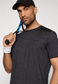 Μαύρο αθλητικό μπλουζάκι κοντομάνικο με υφή, συνδυασμένο με ένα λευκό βραχιολάκι. Ένα μπλε και μαύρο ρακέτα τένις κρατιέται δίπλα από το μοντέλο.