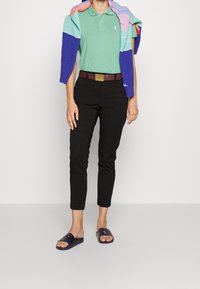 Grön polo skjorta, svarta slim-fit byxor, brun bälte med guld detaljer, flerfärgad stickad tröja över axlarna, marinblå sandaler.