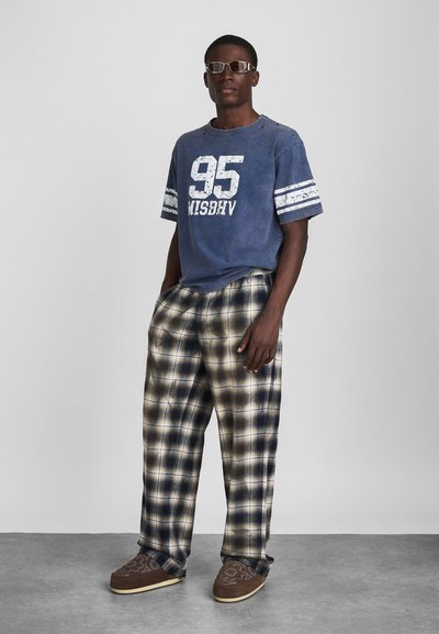 Kék crew-neck póló fehér felirattal és csíkokkal, bézs és tengerészkék kockás nadrággal párosítva. Barna slip-on cipő virágmintával.