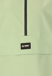 Vihreä vedenpitävä takki, jossa on musta vetoketju, vaakasauma ja suorakulmainen musta etiketti, jossa on valkoisella tekstillä kirjoitettu "SLOPE."