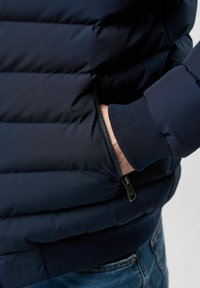 Mano en el bolsillo de una chaqueta de plumas azul marino con cremallera visible y puño de canalé, vistiendo jeans azules.