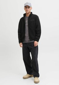 Giacca a zip in pile nera, t-shirt grafica grigia, pantaloni scuri e scarpe beige. Dettagli notevoli: colletto alto, leggera texture sul tessuto.