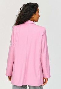 Světlé růžové sako s přiléhavým střihem, hladkou texturou a jedním zadním rozventem; na levém rukávu je našitá nášivka.