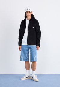 Camisa negra de botones, camiseta blanca, pantalones cortos de mezclilla azules con un patrón de llamas, gorra blanca y zapatillas con suelas texturizadas.