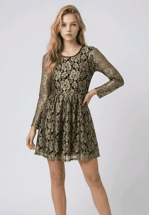 Mujer rubia con vestido de encaje negro de manga larga con patrón floral beige, de pie con una mano en la cadera sobre fondo liso.
