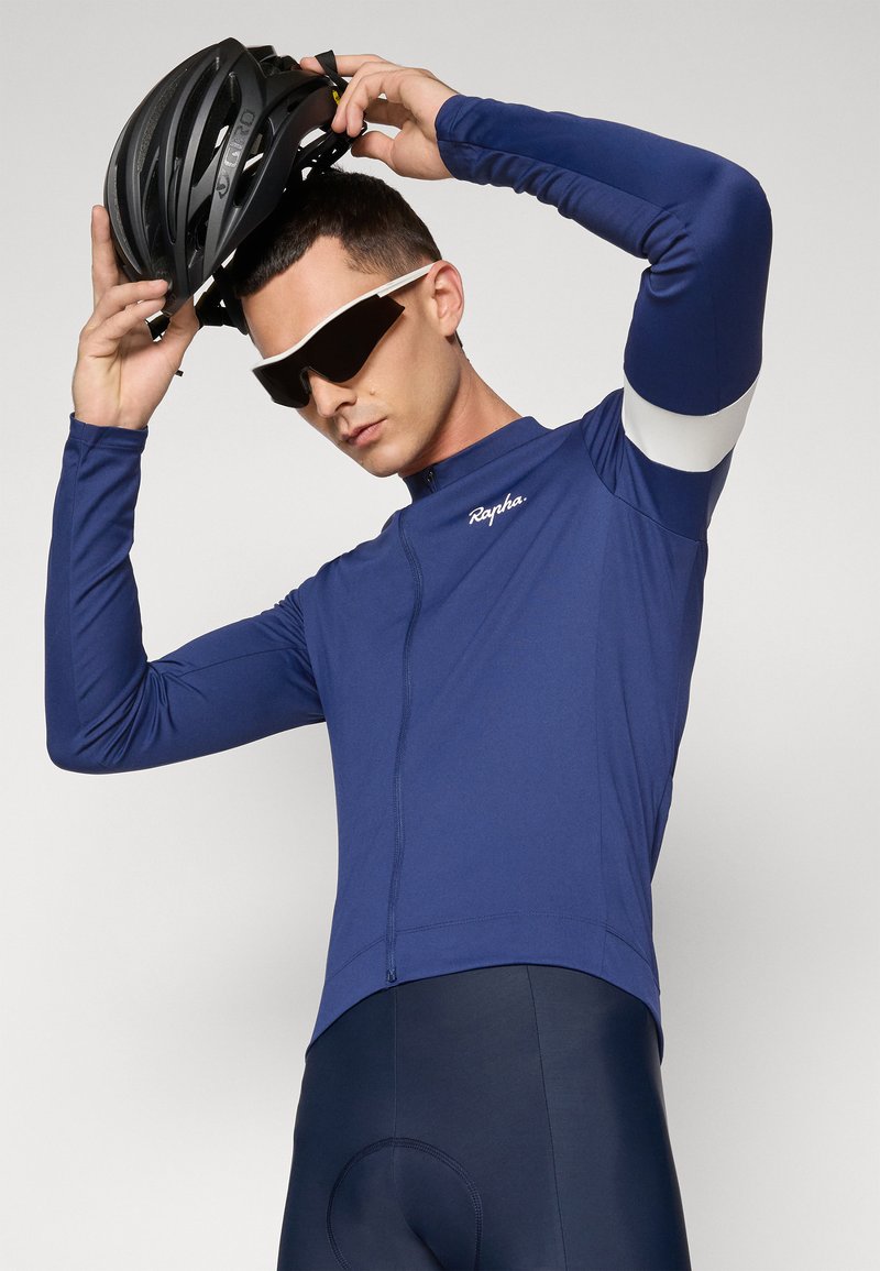 Marineblå cykeltrøje med lange ærmer og lynlås, prydet med hvide detaljer, tekstureret stof og tætsiddende pasform. Modellen bærer en sort hjelm og solbriller.