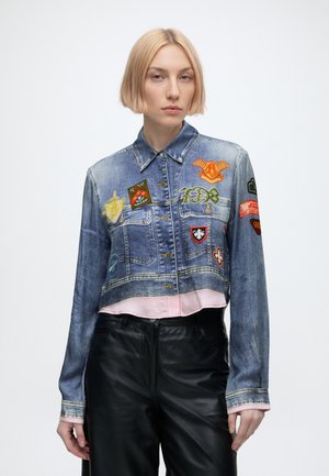 Jeune femme aux cheveux blonds courts porte une veste en jean courte décorée de patchs brodés colorés et un pantalon en cuir noir.