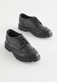 Chaussures habillées en cuir noir avec un design brogue, présentant un bout arrondi, une semelle en caoutchouc épaisse et des perforations détaillées le long des bords.