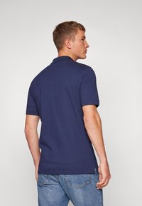 Polo-shirt bleu marine à manches courtes avec un col classique et des fentes latérales ; porté avec un jean bleu clair, mettant en valeur un design simple et épuré.