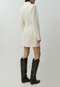 Weißes, maßgeschneidertes Mini-Blazer mit strukturierten Schultern und langen Ärmeln, kombiniert mit schwarzen Lederstiefeln bis zum Knie mit gestickten Mustern.