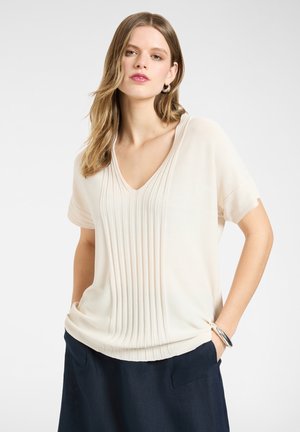 Kobieta w kremowym bluzce z krótkim rękawem, plisowanym dekoltem w kształcie litery V oraz ciemnogranatowych spodniach, stojąca z rękami w kieszeniach na jednolitym jasnym tle.