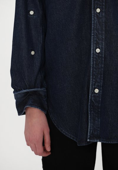 Chemise en denim foncé à boutons blancs, avec manches longues, ourlet arrondi et poignets retroussés. Texture lisse et design décontracté.