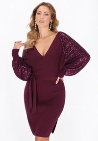 Bordowa dzianinowa sukienka z obszerными rękawami ozdobionymi cekinami, głębokim dekoltem w kształcie litery V i paskiem w talii, z bocznym rozcięciem dla dodatkowego akcentu.