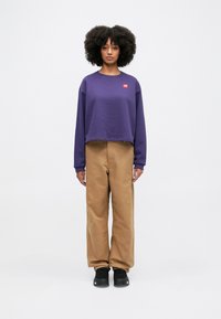Purple cropped sweatshirt with a small red logo, paired with tan wide-leg pants and black sneakers. Smooth fabric, relaxed fit.