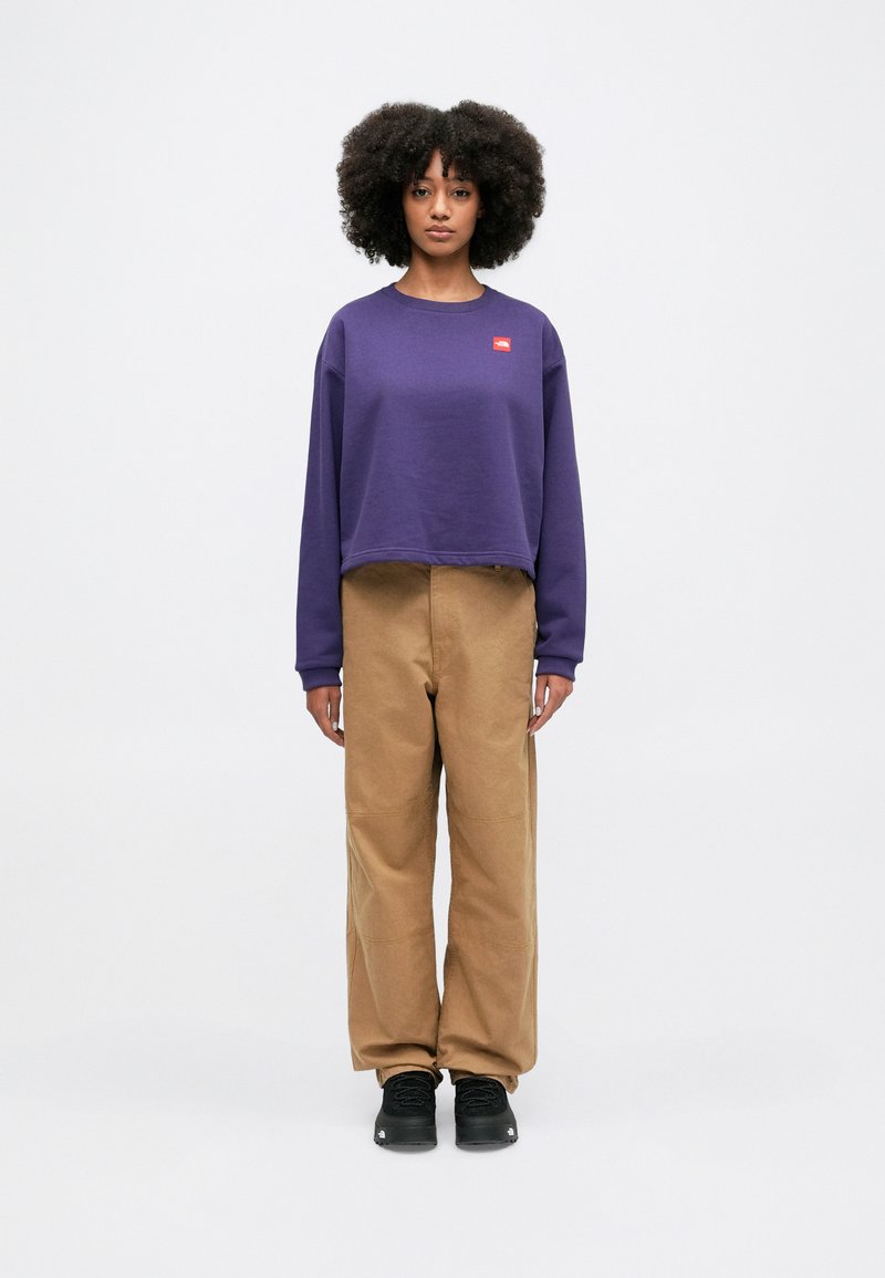 Purple cropped sweatshirt with a small red logo, paired with tan wide-leg pants and black sneakers. Smooth fabric, relaxed fit.