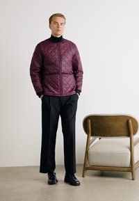 Burgundy quilted jacket with zip closure, black turtleneck, black trousers, and black shoes. Simple design, standing against a light background.