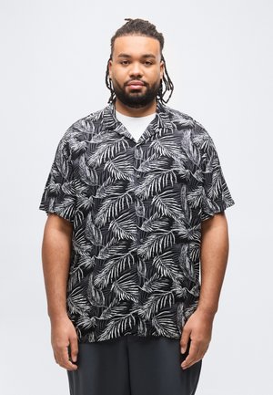 Homme aux cheveux tressés et barbe, portant une chemise noire à manches courtes avec des boutons et un motif de feuilles de palmier blanches, sur un sous-vêtement blanc.