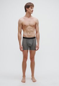 Boxers preto e branco com padrão, feitos de tecido macio, apresentam um cós preto sólido. O design inclui formas geométricas ao longo dos boxers.