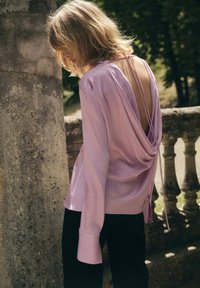 Rosa Satinbluse mit drapierter Rückendetail, langen Ärmeln und breiten Manschetten, kombiniert mit schwarzen Hosen vor einem steinernen Hintergrund.